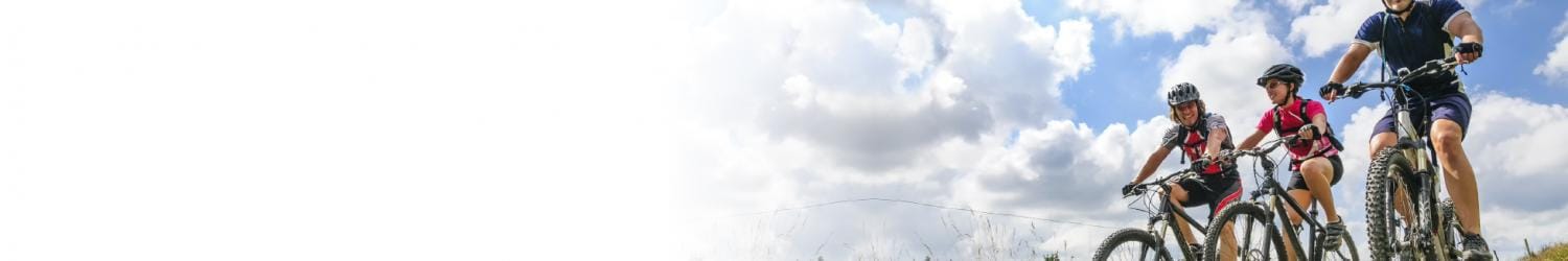 Drei Personen fahren mit Helmen und Fahrradausrüstung unter einem teilweise bewölkten Himmel auf einem Grasweg Rad.