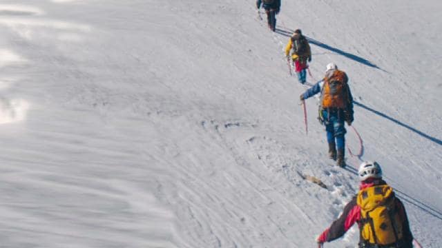 Vier Bergsteiger mit Rucksäcken und Helmen erklimmen einen schneebedeckten Hang. Jeder von ihnen benutzt Trekking-Stöcke und ist durch ein Sicherungsseil verbunden.
