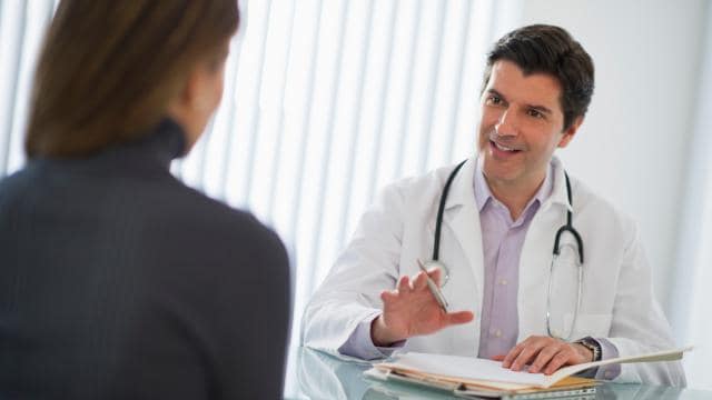 Ein Arzt im weißen Kittel unterhält sich in einem gut beleuchteten Büro an einem Schreibtisch mit einem Patienten.