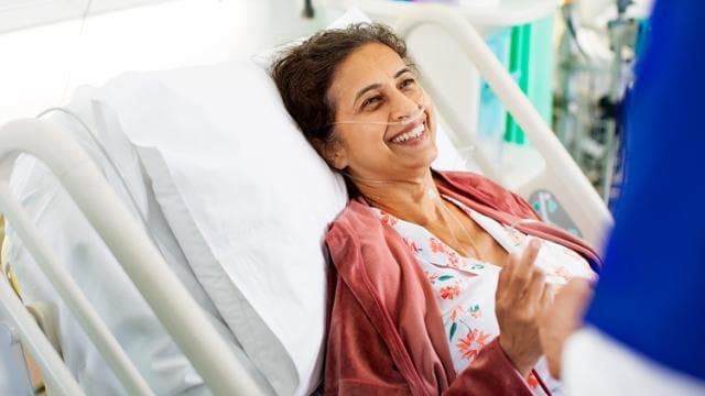 Eine Frau in einem Krankenhausbett lächelt und spricht mit einer Person im blauen OP-Kittel. Sie trägt eine Nasenkanüle und ein geblümtes Kleid unter einer rosa Strickjacke.