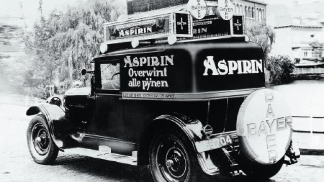 Oldtimer-Lieferwagen mit großen „Aspirin“- und „Bayer“-Logos und einem niederländischen Slogan. Das Fahrzeug ist im Freien geparkt, im Hintergrund sind Bäume und ein Gebäude zu sehen.