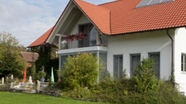Ein zweistöckiges Haus mit rotem Ziegeldach, Solarpanel und Balkon mit Blumen. Der Vorgarten hat einen grünen Rasen und Sträucher.