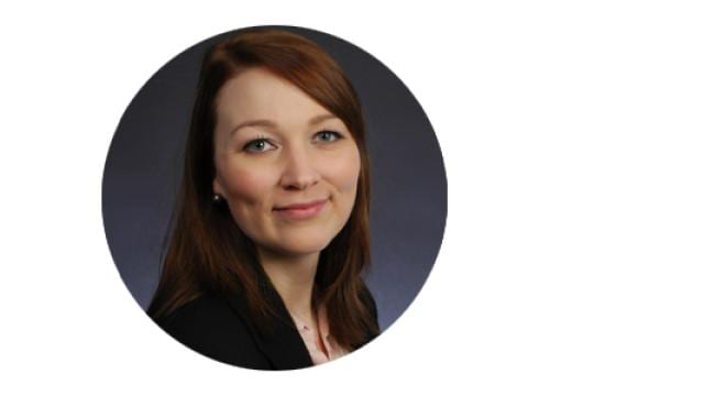 Porträt einer Person mit langen braunen Haaren, die einen schwarzen Blazer trägt und vor einem dunklen Hintergrund leicht lächelt.