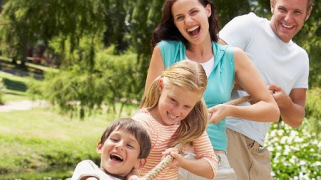 Eine vierköpfige Familie genießt eine Partie Tauziehen im Freien, wobei zwei Kinder und zwei Erwachsene lächelnd an einem Seil zwischen grünen Bäumen und Gras ziehen.