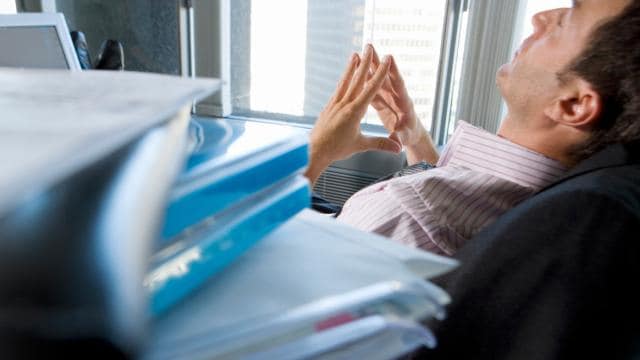 Eine Person lehnt sich in einem Büro mit gefalteten Händen in einem Stuhl zurück und ruht neben einem Stapel Ordner und Papiere.