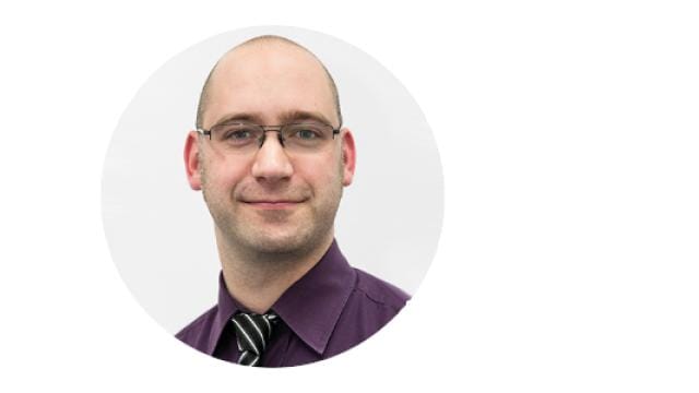 Eine Person mit Brille und Glatze lächelt leicht. Sie trägt ein lila Hemd und eine gestreifte Krawatte vor weißem Hintergrund.