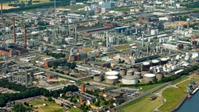 Luftaufnahme eines Industriekomplexes mit zahlreichen großen Gebäuden, Lagertanks und Schornsteinen, umgeben von Grünflächen und einem nahe gelegenen Gewässer.