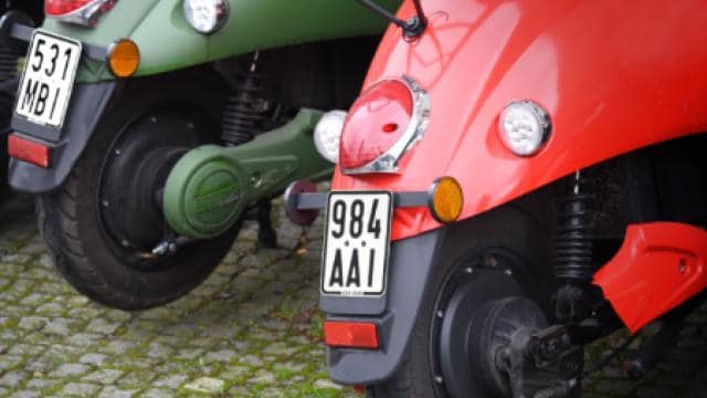 Nahaufnahme von zwei auf Kopfsteinpflaster geparkten Motorrollern. Man sieht deren Heckteile, Reifen und Nummernschilder. Ein Roller ist rot und der andere grün.