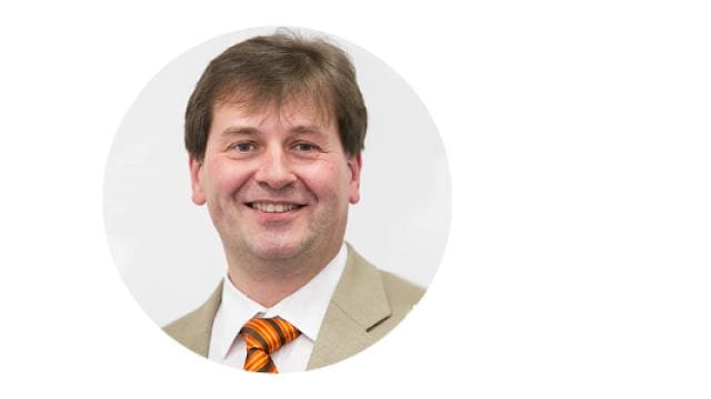 Ein Mann in einem beigen Anzug mit orange gestreifter Krawatte lächelt vor einem weißen Hintergrund in die Kamera.