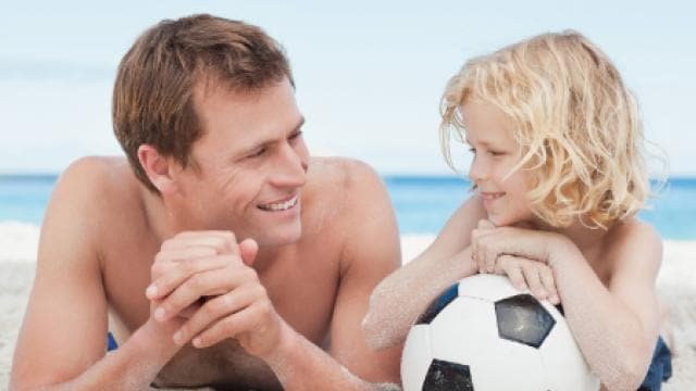 Ein Mann und ein Kind liegen im Sand am Strand und lächeln einander an. Das Kind hat einen Fußball.