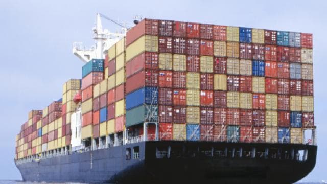 Ein großes Frachtschiff auf See, das zahlreiche gestapelte Frachtcontainer in verschiedenen Farben transportiert. Der Himmel ist klar und das Meer ruhig.