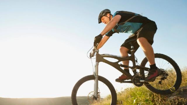 Person, die mit einem Mountainbike einen grasbewachsenen Hügel hinunterfährt, im Hintergrund ein klarer Himmel.