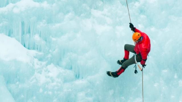 Eine Person in einer roten Jacke und einem orangefarbenen Helm klettert an einer gefrorenen Wand.